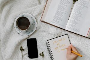 Cena de um momento devocional com Bíblia aberta, caderno com anotações cristãs, caneta e uma xícara de café sobre tecido branco.