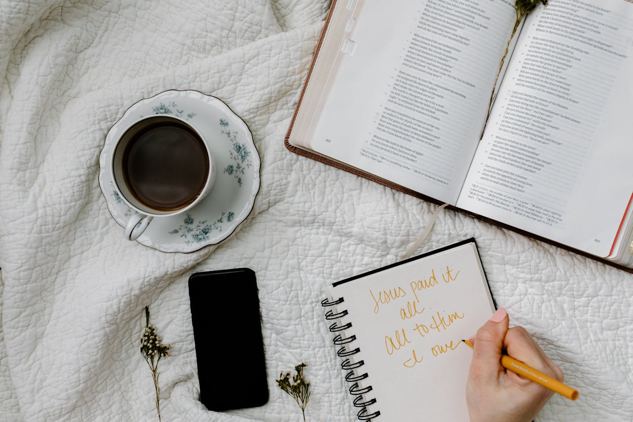 Cena de um momento devocional com Bíblia aberta, caderno com anotações cristãs, caneta e uma xícara de café sobre tecido branco.